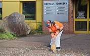 Ausgerüstet mit einem Reisigbesen, einer Warnweste und einer Brille mit runden „30“-Schildern fegt Daniel Kölbl vor dem Tornescher Rathaus Kronkorken zusammen. (Foto: CDU Tornesch)