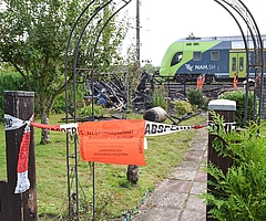 Die Polizei hat den Brandort in der Kleingartenanlage Fuchsberg in Elmshorn abgesperrt. Ein Regionalzug fährt im Schritttempo daran vorbei. Mitarbeiter der Deutschen Bahn untersuchen die technischen Anlagen. (Foto: Frank)